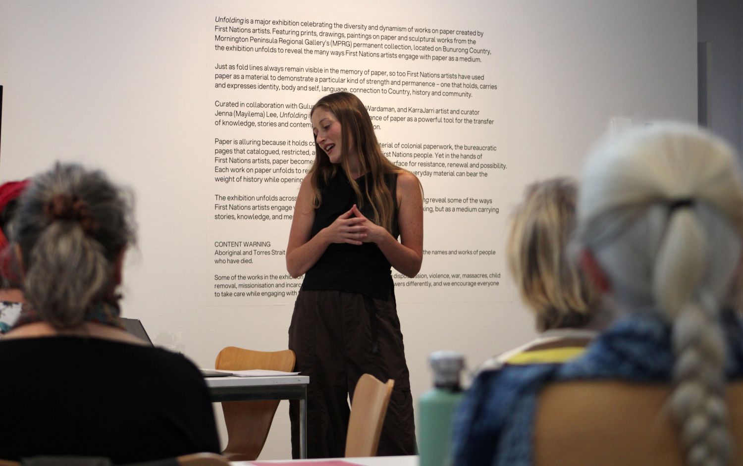 A woman stands at the front of a group of people, speaking to them, with a paragraph of words on the wall behind her